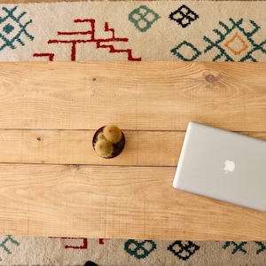 Peut inclure: Un plateau de table en bois avec un petit cactus dans un pot rouge et un ordinateur portable argenté sur le côté droit. La table est posée sur un tapis blanc à motifs géométriques colorés.