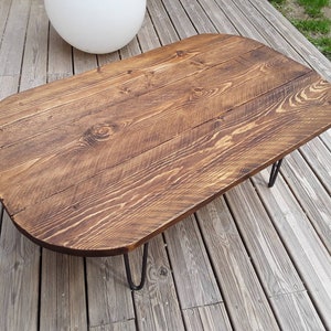 May include: A rustic, oval-shaped coffee table with a dark brown stained wood top. The tabletop is made of wooden planks and rests on black hairpin legs. The table measures approximately 48 inches long and 24 inches wide.
