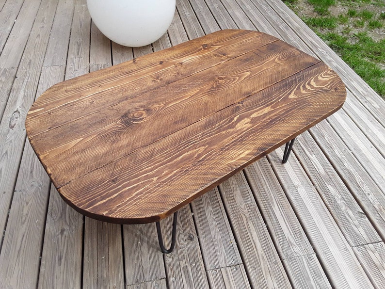 May include: A rustic, oval-shaped coffee table with a dark brown stained wood top. The tabletop is made of wooden planks and rests on black hairpin legs. The table measures approximately 48 inches long and 24 inches wide.