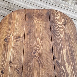 May include: Close-up of a wooden table top with a rounded edge. The table is constructed from several planks of wood, showcasing a rich, warm brown color with visible wood grain and knots. The surface has a rustic, weathered appearance.