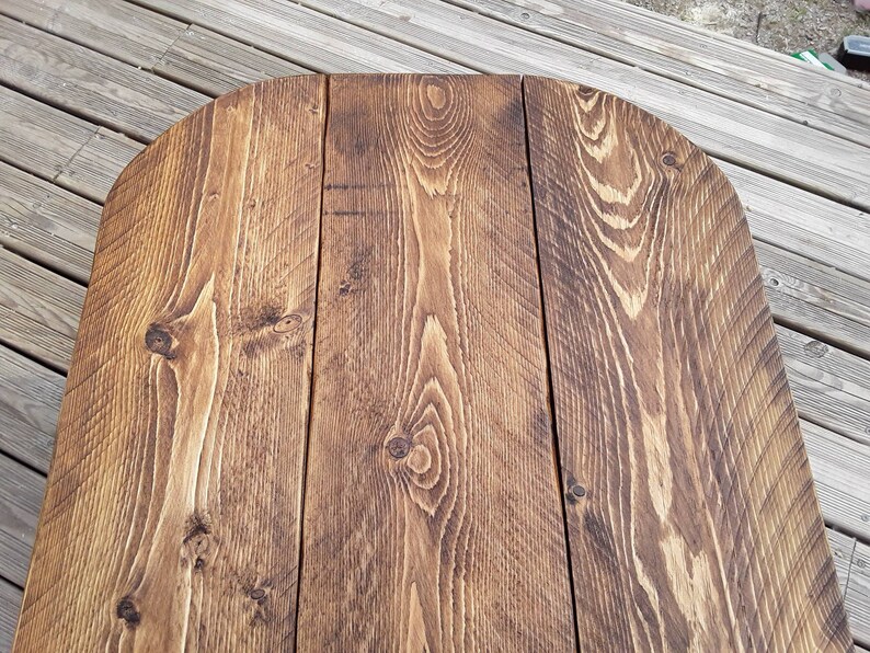 May include: Close-up of a wooden table top with a rounded edge. The table is constructed from several planks of wood, showcasing a rich, warm brown color with visible wood grain and knots. The surface has a rustic, weathered appearance.