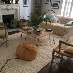 Pu&ograve; includere: Un tappeto bianco a pelo lungo con un motivo a rombi neri, un pouf in pelle marrone, un tavolino da caff&egrave; in legno con una coperta per divano a motivi bianchi e gialli e una sedia bianca con struttura in legno.