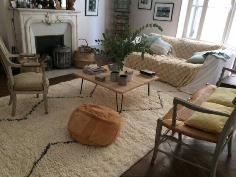 Pu&ograve; includere: Un tappeto bianco a pelo lungo con un motivo a rombi neri, un pouf in pelle marrone, un tavolino da caff&egrave; in legno con una coperta per divano a motivi bianchi e gialli e una sedia bianca con struttura in legno.
