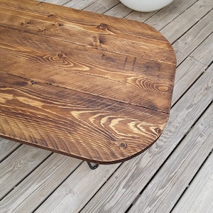 May include: A rustic, wooden coffee table with a rounded edge. The tabletop is made of stained wood with visible grain patterns. The table has dark metal legs and is set on a wooden deck.