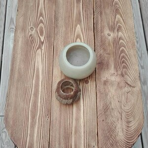May include: A wooden oval-shaped table with a light brown finish. A small, light-colored ceramic bowl and a decorative wooden ring sit on the table. The table is made of wooden planks and has a natural wood grain.