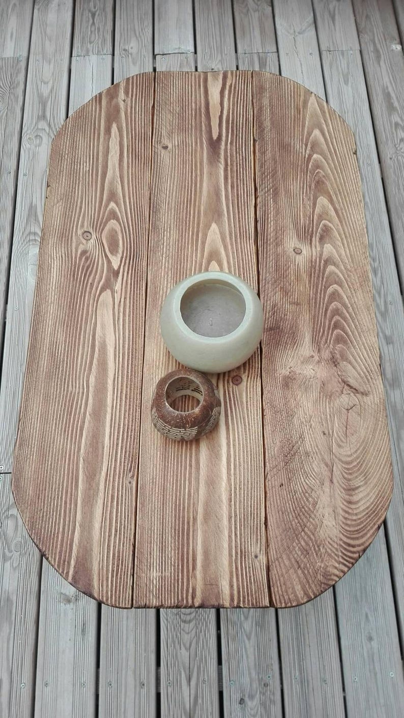 May include: A wooden oval-shaped table with a light brown finish. A small, light-colored ceramic bowl and a decorative wooden ring sit on the table. The table is made of wooden planks and has a natural wood grain.