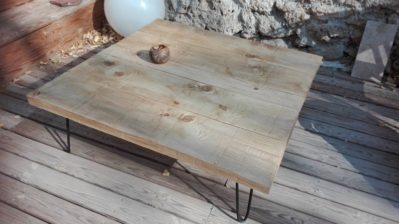 Puede incluir: Una mesa de centro de madera clara con un acabado natural. La mesa tiene una tapa cuadrada y est&aacute; sostenida por cuatro patas de horquilla negras.