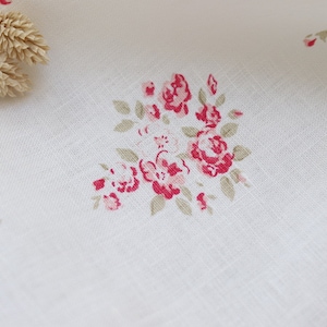 May include: White linen fabric with a delicate floral pattern featuring clusters of red roses and green leaves. The fabric has a subtle texture and is adorned with small sprigs of dried flowers.