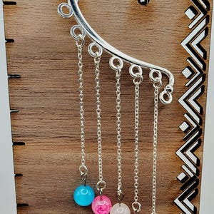 May include: A silver ear cuff with five dangling chains, each ending in a colorful bead. The beads are blue, pink, and white. The ear cuff is displayed on a wooden stand with a decorative geometric pattern.