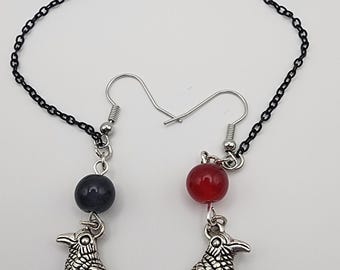 May include: A pair of silver-toned earrings featuring a raven charm, a black chain, and red and black teardrop-shaped beads. The earrings also have a black bead and a red bead. The raven charms are detailed with etched feathers.