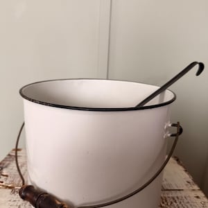 May include: White enamel bucket with a black rim and a metal handle. The handle has a wooden grip. A black metal ladle rests inside the bucket. The bucket sits on a distressed wooden surface.