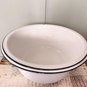 May include: A white enamelware bowl with a black rim. The bowl has a classic, rounded shape and is set against a rustic, distressed wooden surface. The interior of the bowl is clean and bright.