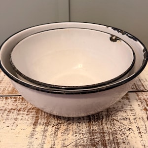 May include: Two white enamelware bowls nested together. The bowls have a black rim and show signs of wear, with some areas of the enamel chipped away. The bowls are set on a distressed, light-colored wooden surface.