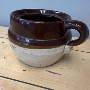 May include: A brown and white ceramic crock with a handle. The crock has a dark brown glazed top and a lighter brown band, with a white base. The handle is curved and attached to the side. The crock is sitting on a wooden surface.