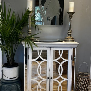 May include: White mirrored cabinet with two doors and a distressed finish. The doors have a decorative lattice design. The cabinet is sitting on a wooden floor and has a small plant in a white pot to the left.
