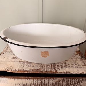 May include: A white enamelware bowl with a black rim. The bowl has a slightly distressed appearance, with some wear and tear visible. A faded sticker is on the side of the bowl. The bowl is sitting on a rustic wooden surface.