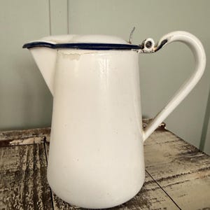 May include: White enamel pitcher with a curved handle and lid. The pitcher has a blue trim around the lid and shows signs of wear. The pitcher is sitting on a distressed wooden surface. A vintage kitchenware item.