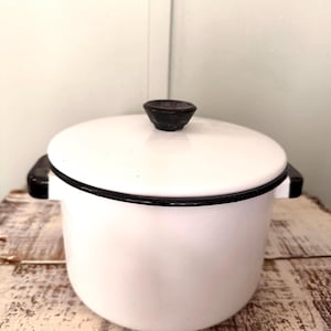 May include: White enamel pot with a black rim and handles. The pot has a matching lid with a black knob. The pot is sitting on a distressed wooden surface. A classic kitchenware item.