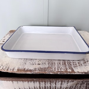 May include: A white enamel baking pan with a blue rim. The rectangular pan is empty and sits on a distressed wooden surface. The pan is a classic kitchenware item.