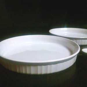 Op de afbeelding: Twee witte keramische bakvormen, waarvan er één iets groter is dan de andere, met geribbelde randen. De vormen zijn leeg en lijken nieuw te zijn.