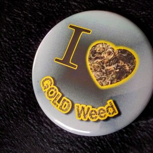 May include: A round button with a gray background and a yellow heart filled with a brown and green image. The text "I" is in black and the text "Gold Weed" is in yellow.