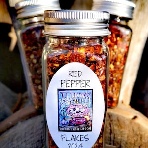 May include: Three glass jars with silver lids filled with red pepper flakes. The center jar has a white oval label that reads "Red Pepper" and "Allicin's Ranch" with a cartoon image of a red truck and garlic. The label also says "Flakes 2024" and "allicinsranch.com".