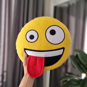 Può includere: Cuscino emoji in peluche giallo con un grande sorriso e una lingua rossa che sporge. L'emoji ha grandi occhi bianchi con pupille nere.