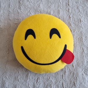 Peut inclure: Oreiller emoji en peluche jaune avec un grand sourire et une langue rouge qui sort.
