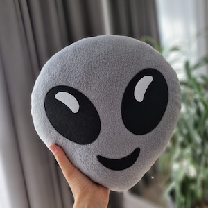 May include: A grey alien emoji plush pillow with black eyes and a smiling mouth.