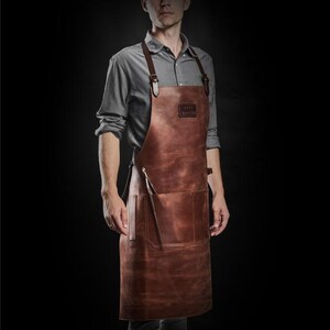 May include: A brown leather apron with adjustable straps and a large pocket. The apron has the text "GREY +  KNOB" printed on the front.