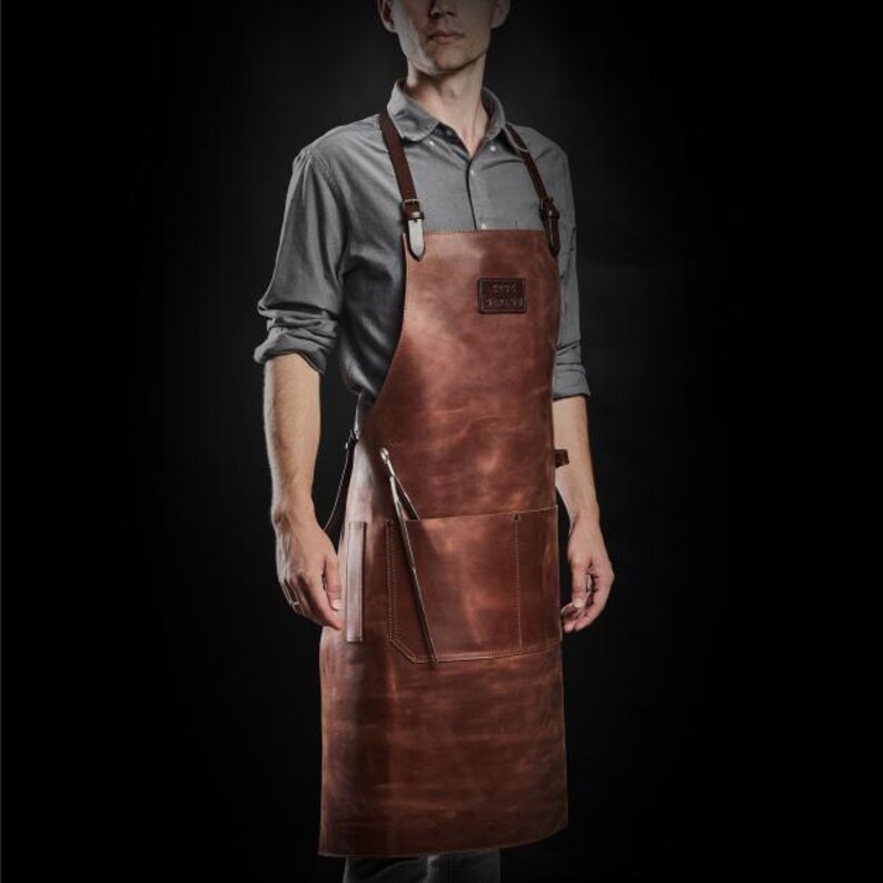 May include: A brown leather apron with adjustable straps and a large pocket. The apron has the text "GREY +  KNOB" printed on the front.