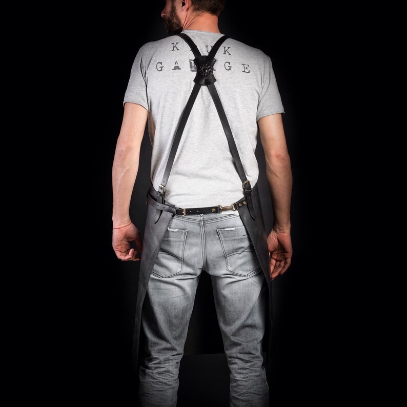 May include: Black leather apron with suspenders, worn over a gray t-shirt and jeans. The apron has a belt and brass hardware. The t-shirt has the text "KUK GARAGE" printed on the back.
