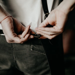 May include: A person adjusts the straps of a pair of black suspenders. The suspenders are attached to a pair of dark green pants.