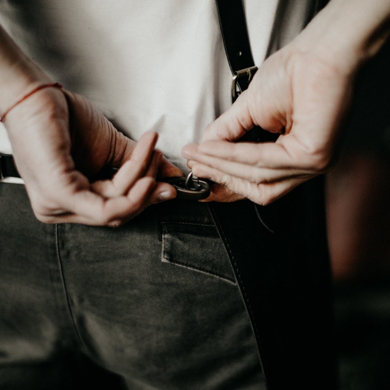 May include: A person adjusts the straps of a pair of black suspenders. The suspenders are attached to a pair of dark green pants.