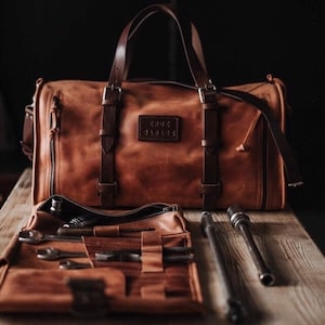 Könnte beinhalten: Eine braune Ledertasche mit Reißverschluss und einem Lederriemen. Die Tasche hat ein Etikett mit der Aufschrift "Cross Garage". Die Tasche ist offen und ein brauner, zusammenrollbarer Werkzeughalter aus Leder mit Werkzeugen ist sichtbar. Zwei Metallwerkzeuge liegen auf der Holzoberfläche.