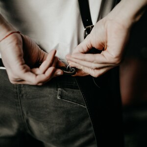 May include: Close-up of a person adjusting black leather suspenders attached to dark gray pants. The person is wearing a white t-shirt. A red bracelet is visible on the left wrist. The background is dark.