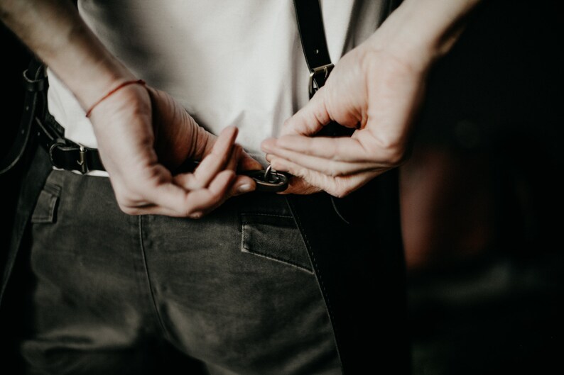 May include: Close-up of a person adjusting black leather suspenders attached to dark gray pants. The person is wearing a white t-shirt. A red bracelet is visible on the left wrist. The background is dark.