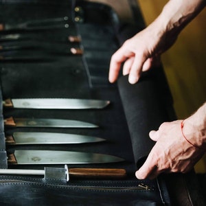 May include: A black leather knife roll with a zipper closure. The roll is open and contains a set of knives with wooden handles. The roll is labeled "KRUK" and has a stitched design.
