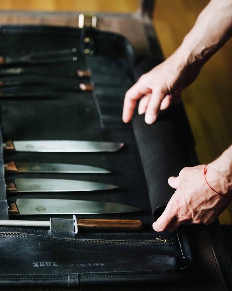 May include: A black leather knife roll with a zipper closure. The roll is open and contains a set of knives with wooden handles. The roll is labeled "KRUK" and has a stitched design.