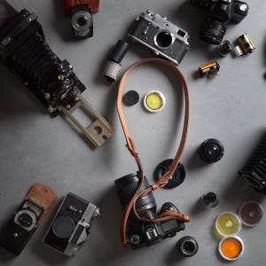 May include: A collection of vintage cameras, film canisters, and camera straps on a gray surface. The cameras include a black and silver rangefinder camera, a black and silver SLR camera, and a black bellows camera. The film canisters are various sizes and colors. The camera straps are brown leather.