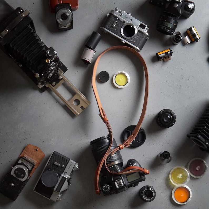 May include: A collection of vintage cameras, film canisters, and camera straps on a gray surface. The cameras include a black and silver rangefinder camera, a black and silver SLR camera, and a black bellows camera. The film canisters are various sizes and colors. The camera straps are brown leather.
