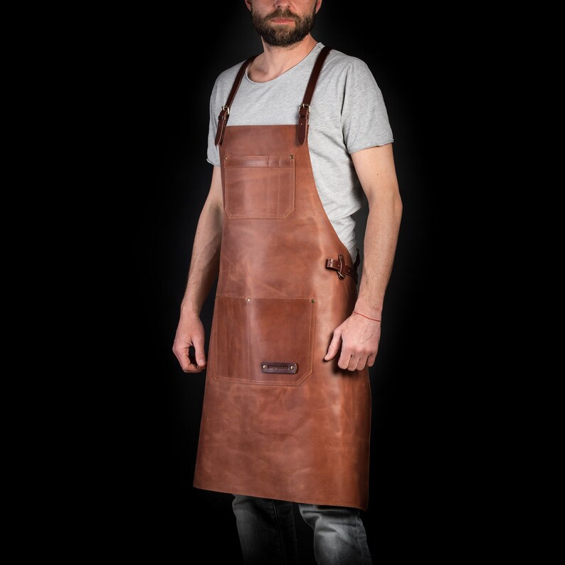 May include: A brown leather apron with two large pockets and adjustable straps. The apron has a rectangular patch with text on the lower pocket and is worn over a gray t-shirt. The apron is designed for use in the kitchen or workshop.