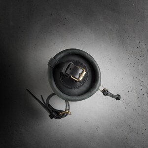May include: A dark gray leather cylindrical bag with a black leather strap and a gold buckle. The bag is on a gray textured surface.