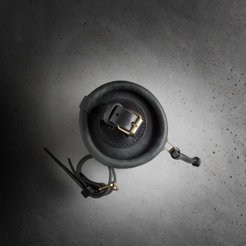 May include: A dark gray leather cylindrical bag with a black leather strap and a gold buckle. The bag is on a gray textured surface.