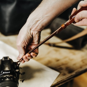 May include: A person attaches a brown leather camera strap to a black Nikon DSLR camera.