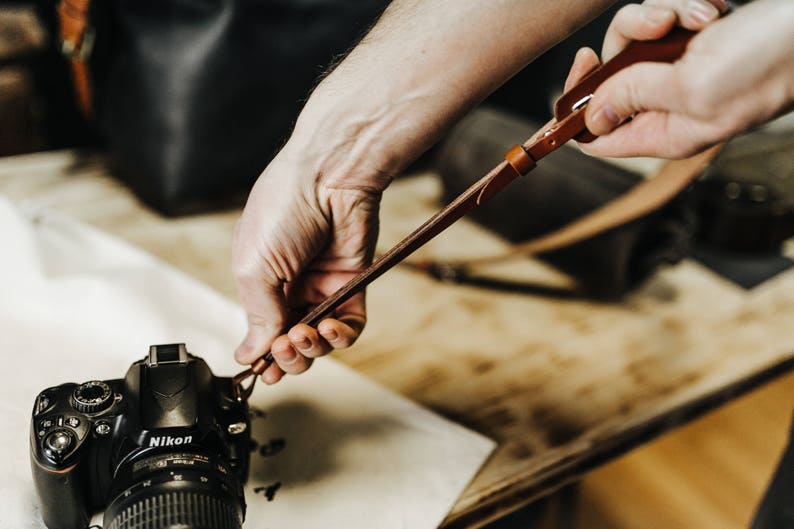 May include: A person attaches a brown leather camera strap to a black Nikon DSLR camera.