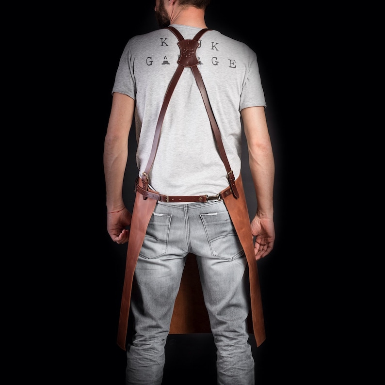 May include: A brown leather apron with adjustable straps and a buckle closure. The apron has a large pocket on the front and the text "KUK GARAGE" printed on the back.