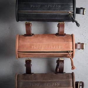 May include: Three leather handlebar bags in black, brown, and tan with the text "KRUK GARAGE" embossed on each. The bags have a zipper closure and adjustable straps.