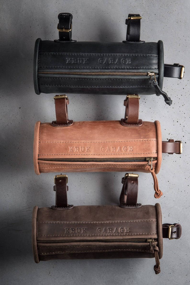 May include: Three leather handlebar bags in black, brown, and tan with the text "KRUK GARAGE" embossed on each. The bags have a zipper closure and adjustable straps.