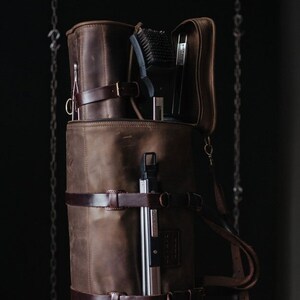 May include: A brown leather bag with a zippered top and multiple straps. The bag is filled with various tools, including a brush, a metal tool, and a long, thin metal tool.